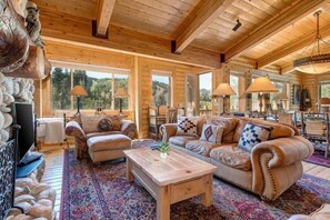 A log cabin interior with a wooden ceiling, large windows, a sofa set, and a patterned rug.