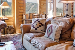 Interior de una cabaña de troncos con un sofá de cuero, cojines estampados, una lámpara y un interior rústico de madera.