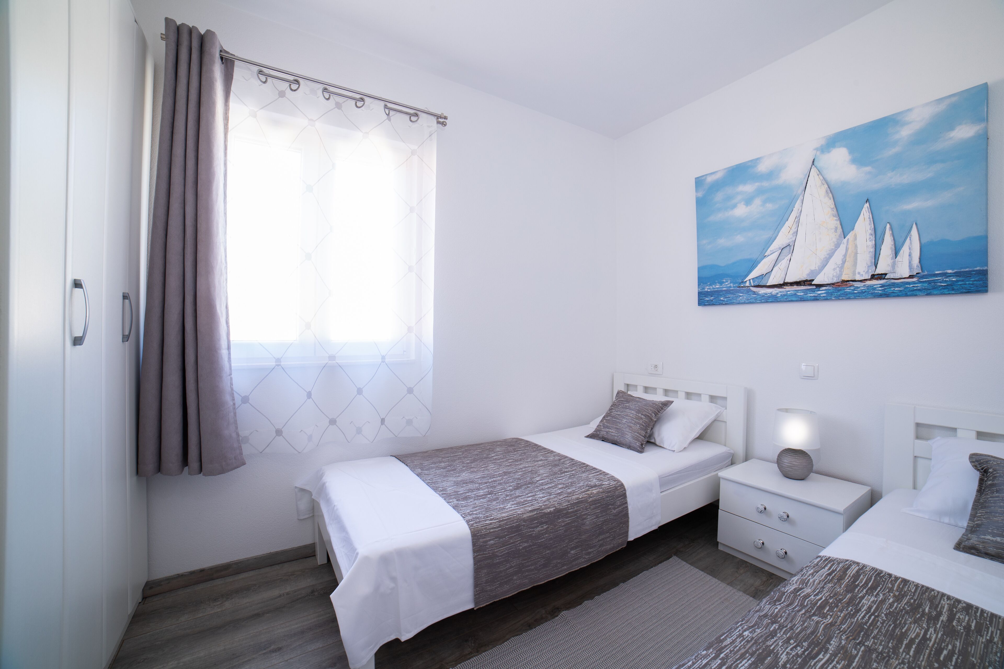 A bedroom with two beds, a painting of sailboats, a window with curtains, and a nightstand with a lamp.