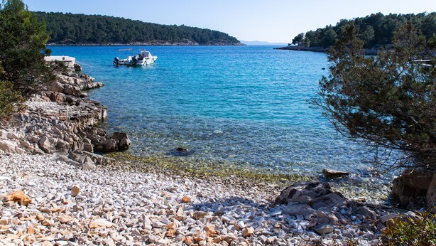 Una costa rocciosa con acque cristalline blu, una barca ancorata a riva e una collina boscosa sullo sfondo.