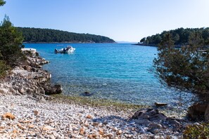 A rocky shoreline with clear blue water, a boat anchored near the shore, and a forested hill in the background.