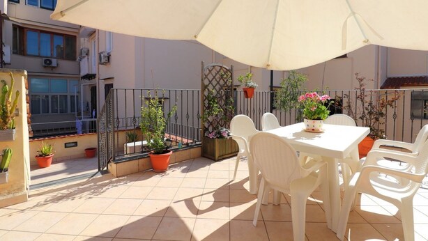 An outdoor patio with a white table and chairs, surrounded by potted plants and a large umbrella.