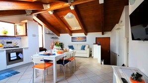 A modern kitchen with a dining area, wooden ceiling beams, a TV mounted on the wall, and a painting above the couch.