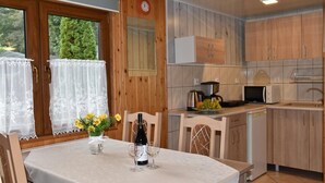 A wooden interior with a dining table set for two, a kitchen with wooden cabinets, and a window with lace curtains. - Bungalows an der Ostsee (Kołobrzeg)