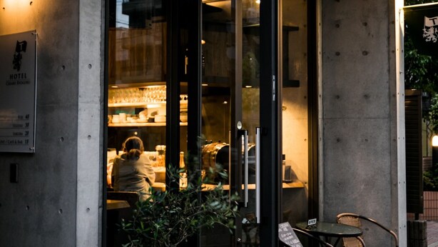 L’entrée d’un hôtel, avec une porte en verre, un pot de fleurs et un petit espace extérieur aménagé avec des sièges.