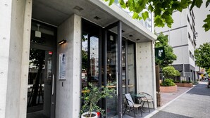 L’extérieur d’un bâtiment moderne, avec une entrée dotée d’une porte en verre, un pot de fleurs et un petit espace extérieur aménagé avec des chaises.
