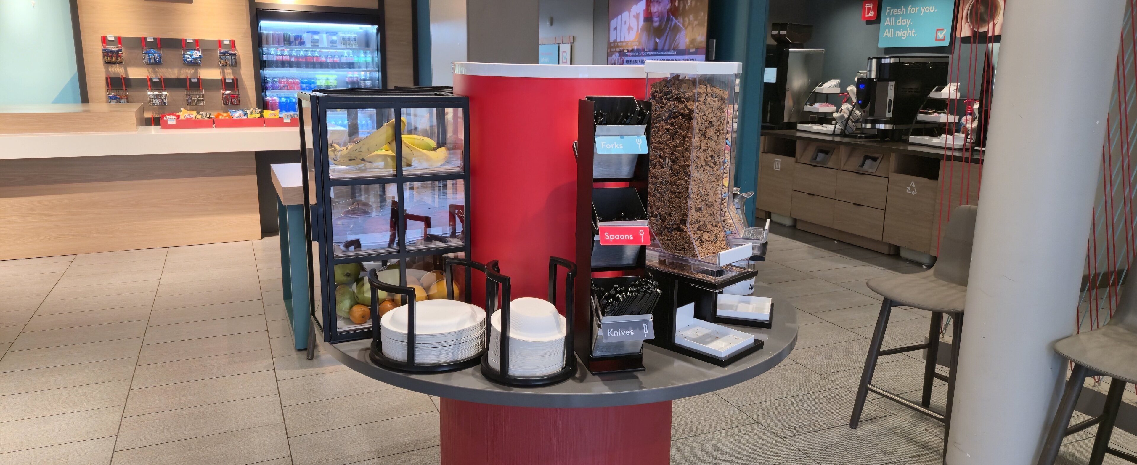 A self-service food station with a glass display case containing bananas, a bowl of fruit, and a stack of white plates.