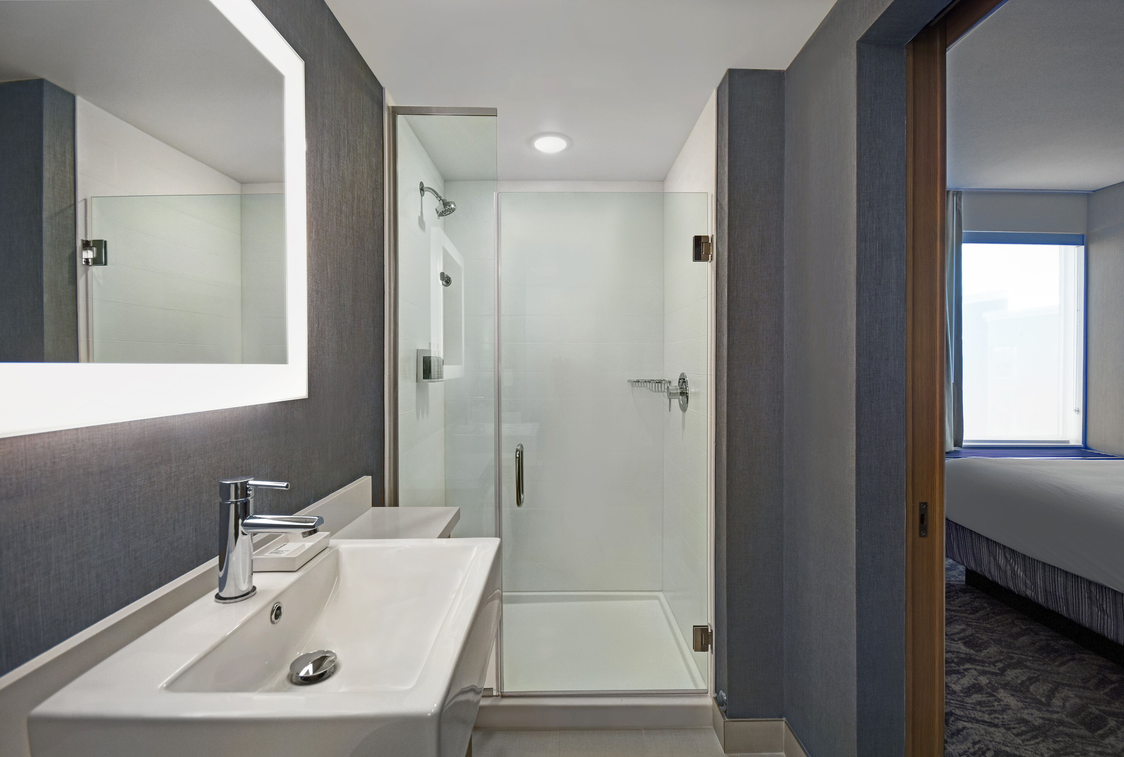 A modern bathroom with two sinks, a glass shower enclosure, and a visible bedroom with a bed and window.