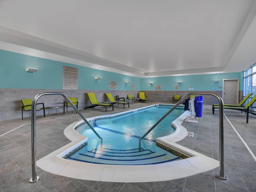 An indoor swimming pool with clear pool rules signs, green lounge chairs, and a tiled floor.