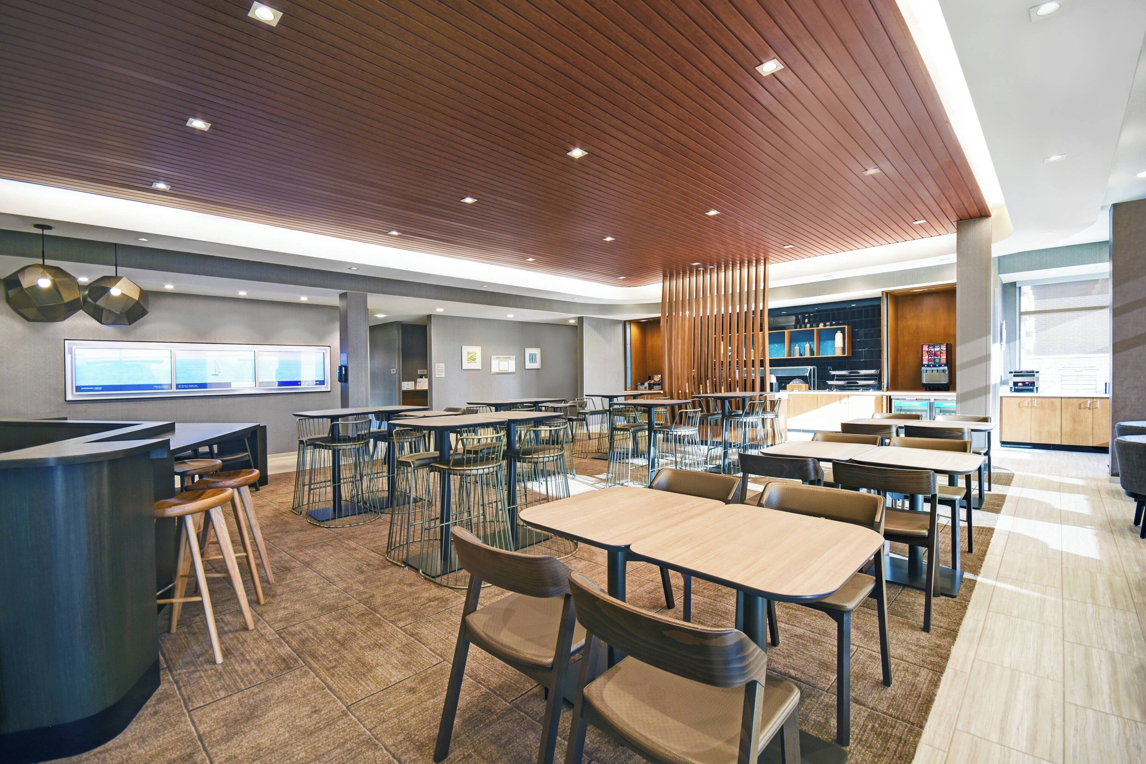 A modern dining area with multiple tables and chairs, a kitchen in the background, and a wooden ceiling.