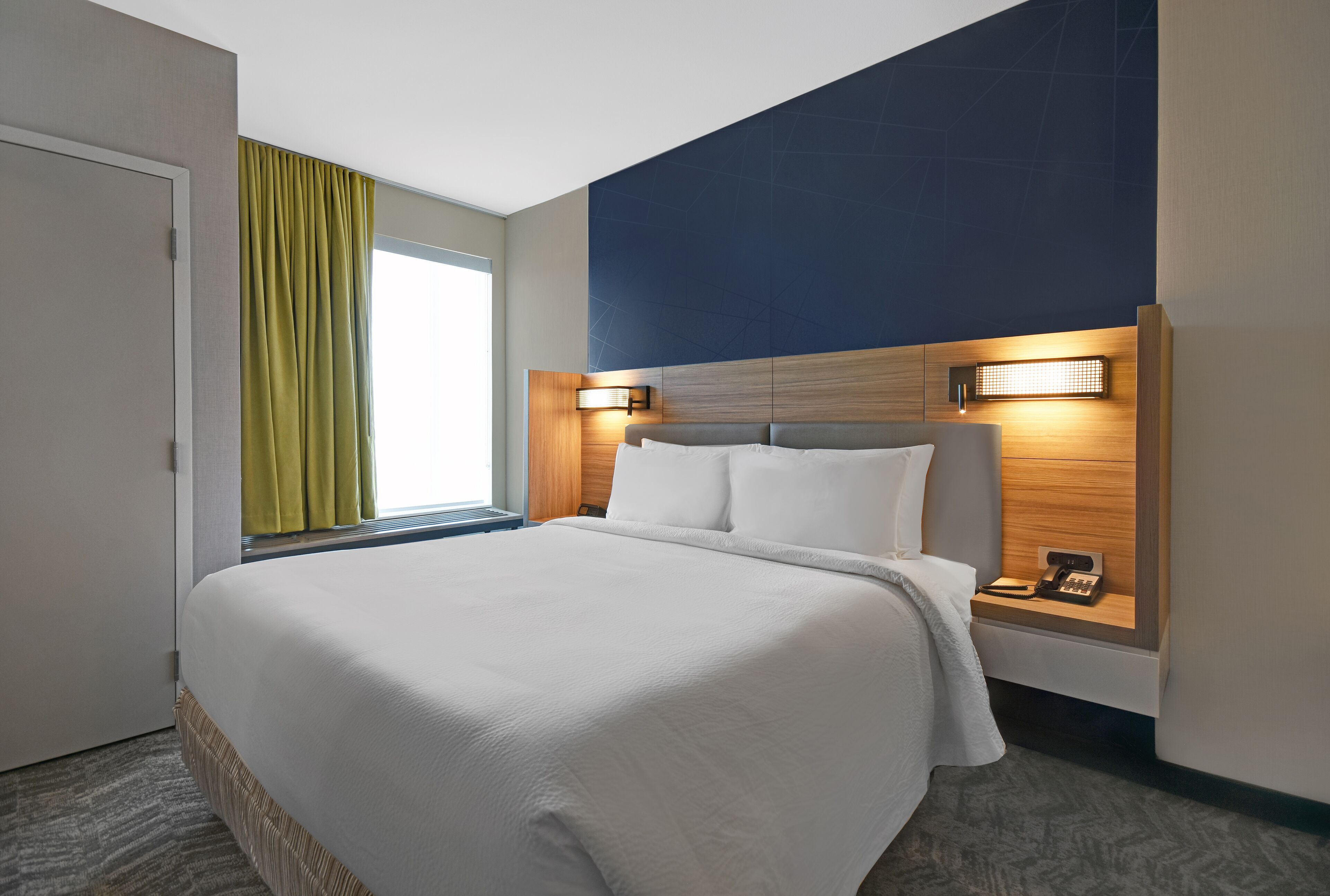A hotel room with a large bed, a nightstand with a phone, and a window with curtains.