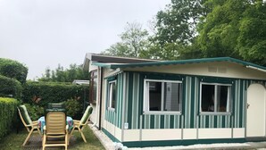 A small, single-story mobile home with a green and white exterior, a front porch with a table and chairs, and a small garden area. - 5-Star Park Texel (De Cocksdorp)