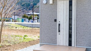 A modern building with a grey brick facade, a wooden door with a vertical metal grille, and a small outdoor area with a tree and a gravel path. - Bon Moment B (Fujikawaguchiko)