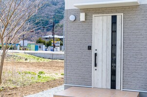 Um edifício moderno com fachada em tijolos cinzas, uma porta de madeira com grade metálica vertical e uma pequena área externa com uma árvore e um caminho de cascalho.