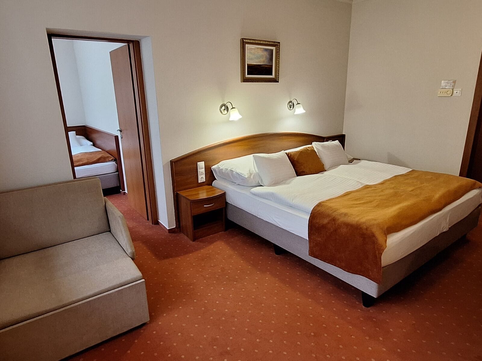 A hotel room with a bed, a nightstand, a lamp, a picture on the wall, and a door leading to another room.