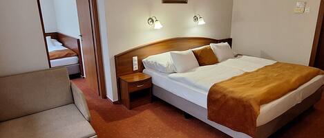 A hotel room with a bed, a nightstand, a lamp, a picture on the wall, and a door leading to another room.