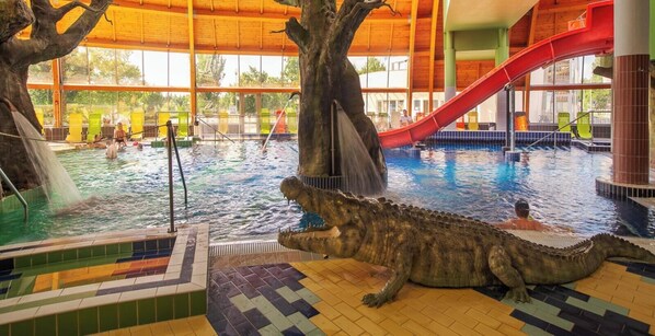 Una piscina interior con una gran escultura de caimán, un tobogán rojo y una estructura con forma de árbol.