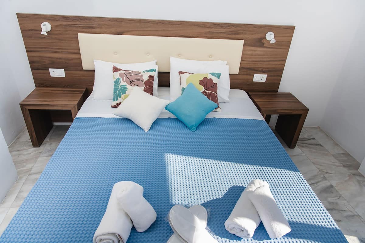 Una cama bien tendida con ropa de cama azul, dos almohadas con estampados florales y un par de toallas enrolladas en el suelo.