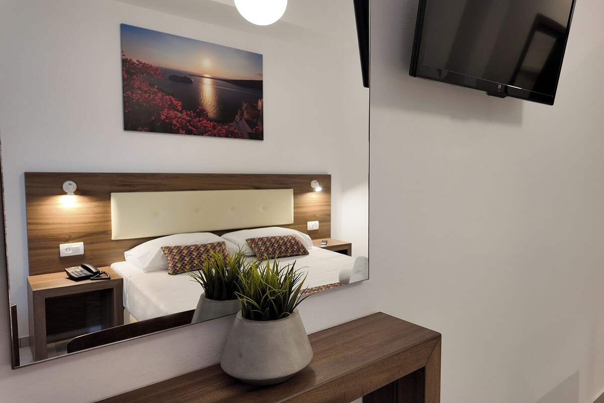 Una moderna camera d'albergo con un ampio letto, comodini, uno specchio, una TV a schermo piatto e un quadro raffigurante un tramonto sull'acqua.