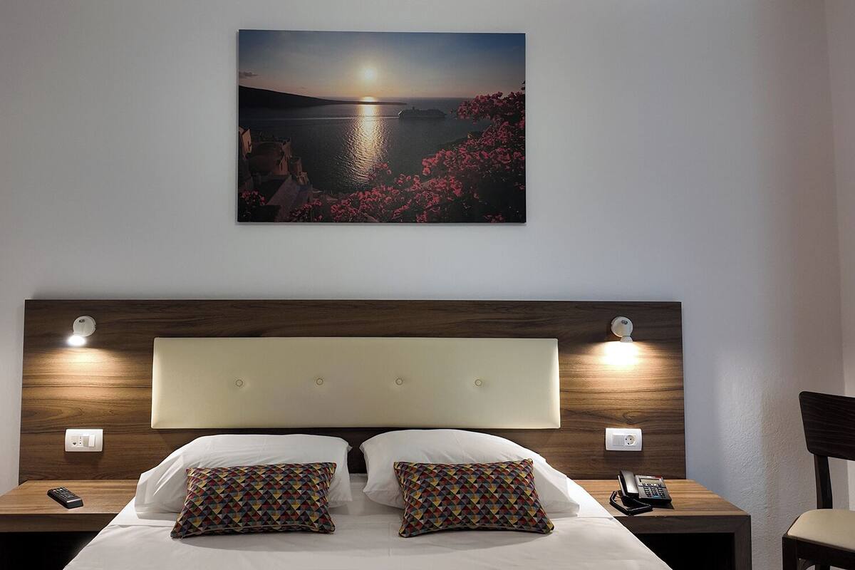 Habitación de hotel con una cama grande, dos almohadas, una mesita de noche con teléfono y un cuadro de un atardecer sobre el agua en la pared.
