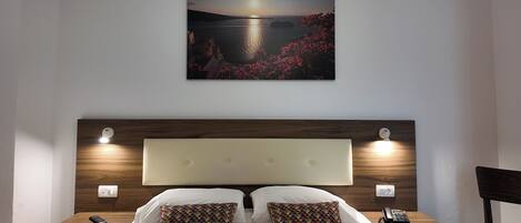 A hotel room with a large bed, two pillows, a nightstand with a phone, and a painting of a sunset over water on the wall.