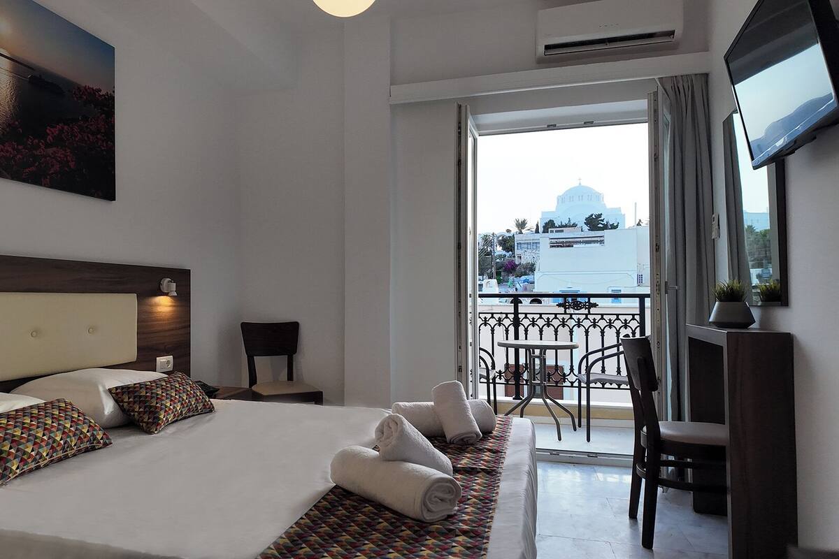 Habitación de hotel con una cama grande, un cuadro en la pared, un balcón con vistas a un edificio y un televisor montado en la pared.