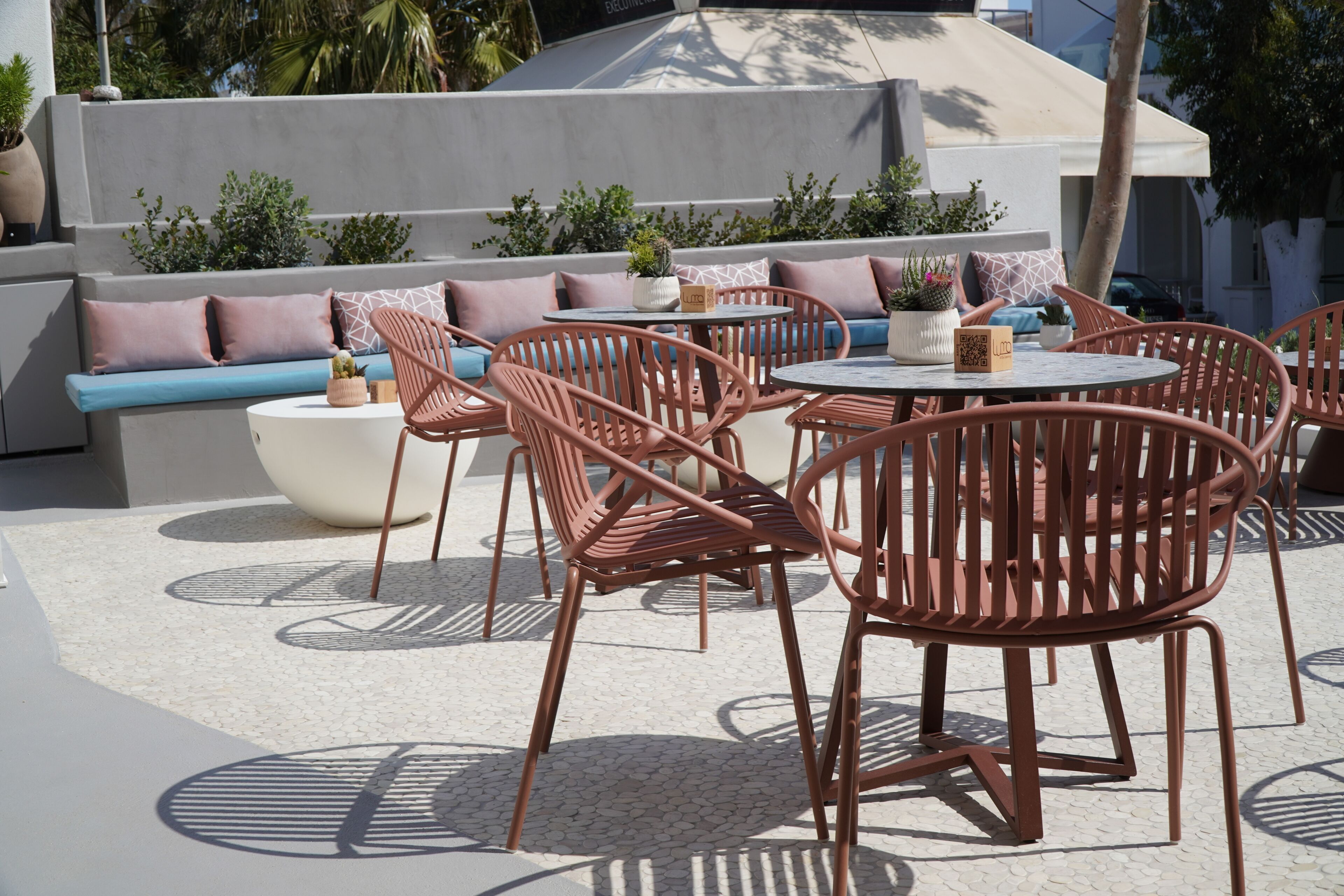 An outdoor seating area with round tables and chairs, a cushioned bench, and potted plants.