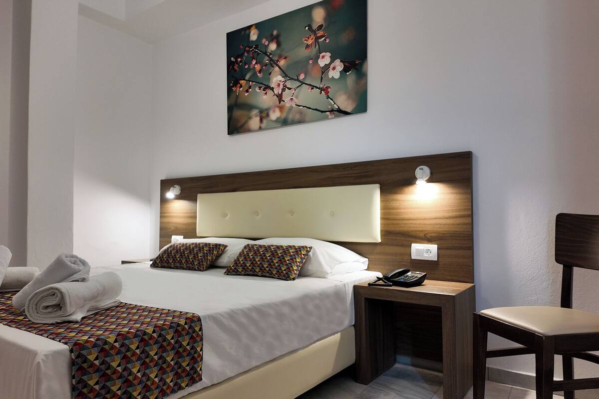 A hotel room with a bed, a bedside table with a phone, a chair, and a painting on the wall.