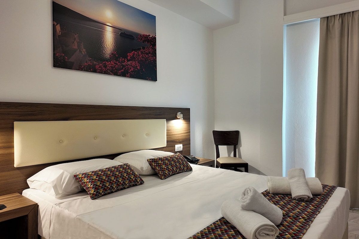 A hotel room with a large bed, a headboard, a nightstand, a chair, a window with curtains, and a painting on the wall.