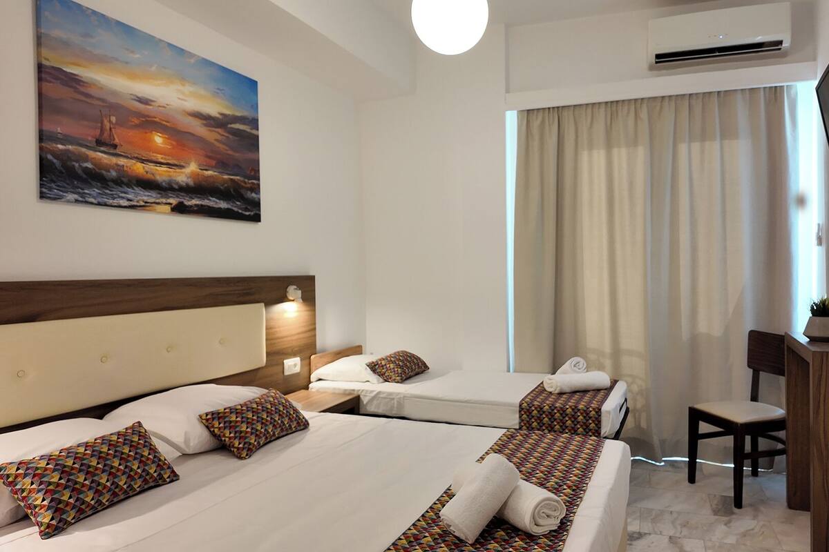 A hotel room with two beds, a TV, a desk, and a painting of a sunset over the ocean.