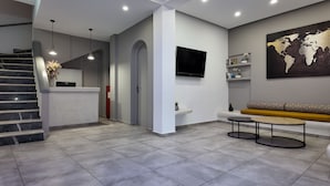 A modern hotel lobby with a reception desk, a staircase, a seating area with a sofa and coffee table, a flat-screen TV mounted on the wall, and a world map artwork. - Hotel Santorini (Santorini)