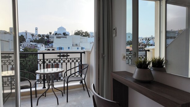 A balcony with a table and chairs, a potted plant, and a view of white buildings and a church. - Hotel Santorini (Santorini)