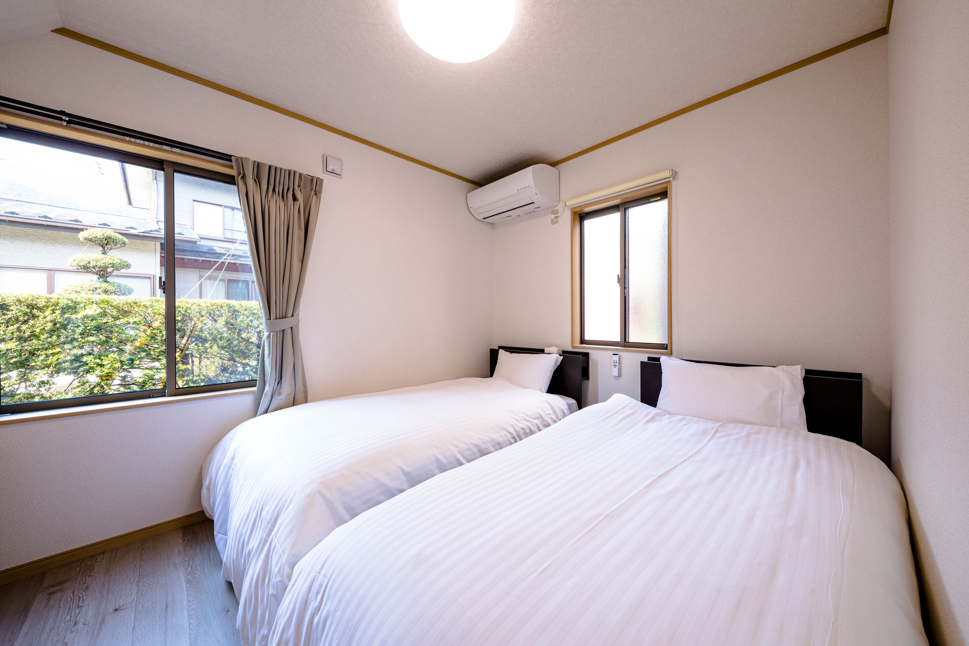 Ett hotellrum med två sängar, ett fönster med gardiner, en luftkonditioneringsanläggning och utsikt över grönområden utanför.