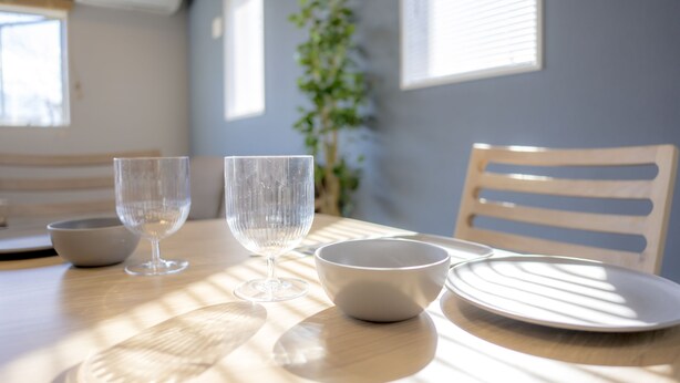 Uma mesa de jantar com uma tigela, dois copos de vinho e um prato, em um ambiente com janela e planta.
