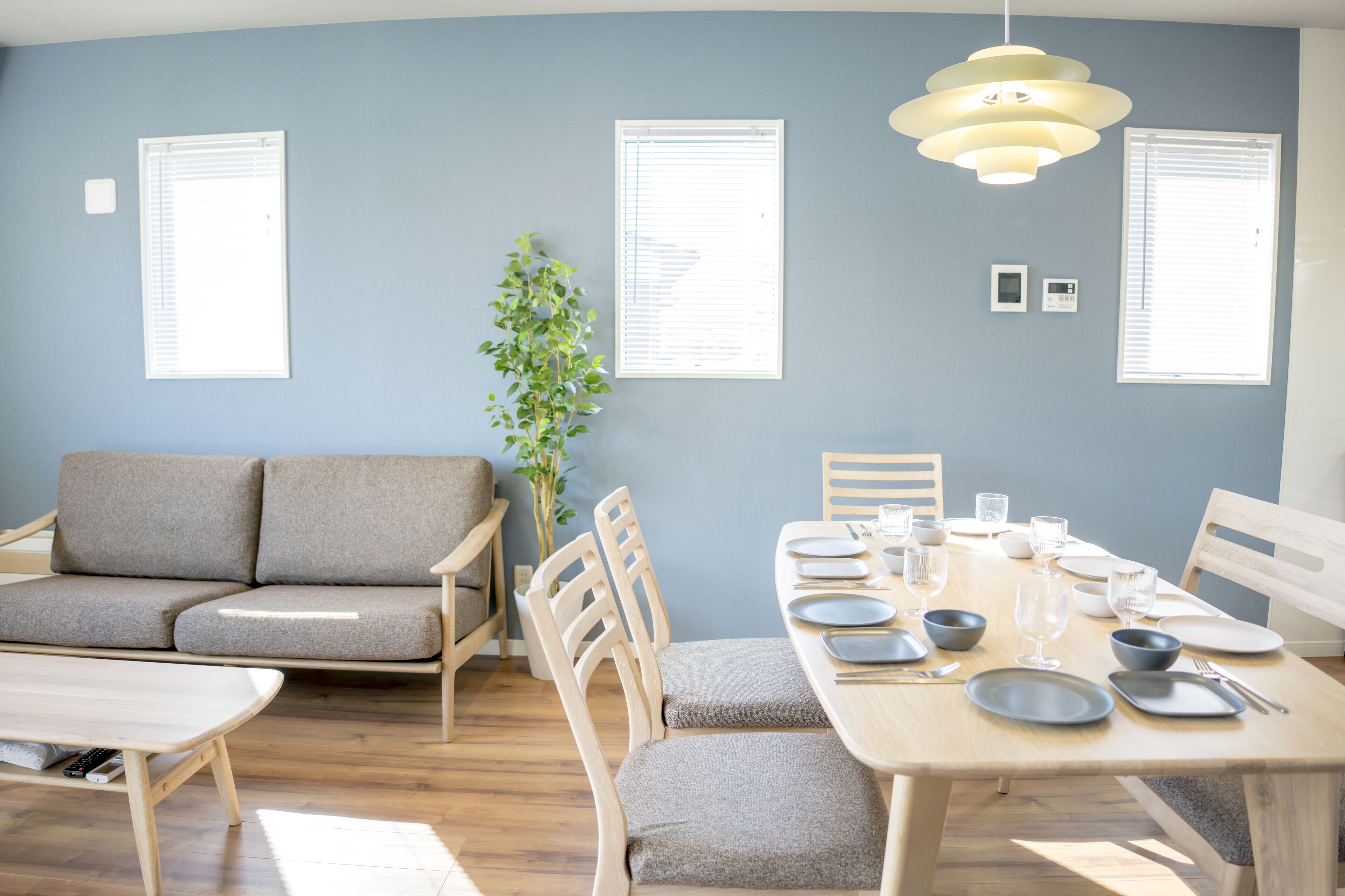 A modern living room with a sofa, dining table set for four, and a large window with blinds.