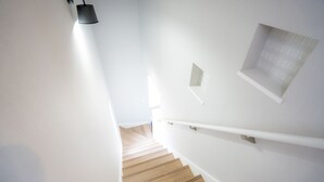 Escada com degraus de madeira, paredes brancas e luminária preta fixada na parede.