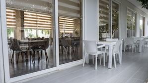 A modern outdoor dining area with glass walls, wooden blinds, and a mix of dark and white chairs. - Side Spring Hotel (Manavgat)