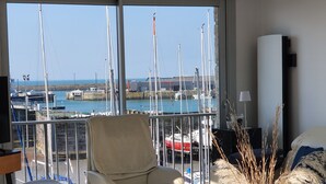 A modern living room with a large window overlooking a marina. There are several chairs and a table with a vase of dried plants.