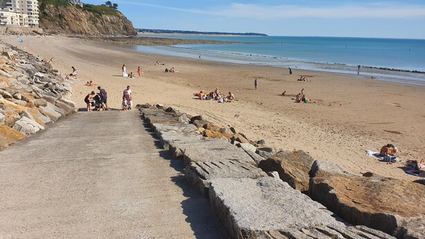 A beach with a stone pathway leading to the shore, people sunbathing, and buildings in the background.