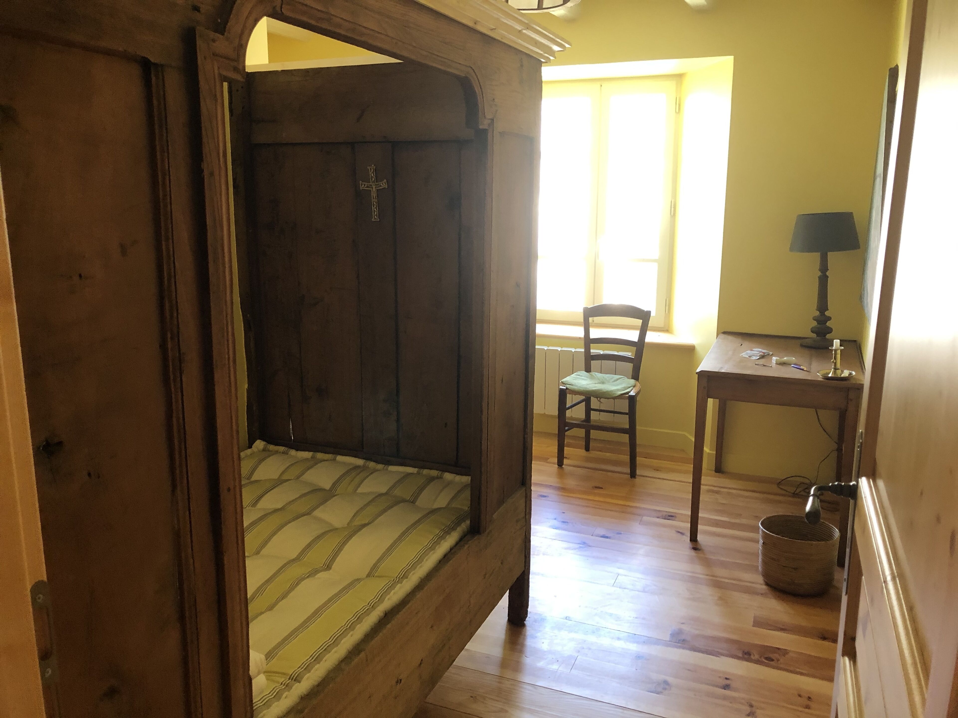 A wooden bed with a cross on the headboard, a chair, a small table with a lamp, and a window with blinds.