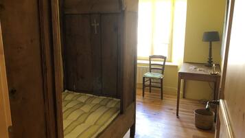 A wooden bed with a cross on the headboard, a chair, a small table with a lamp, and a window with blinds.