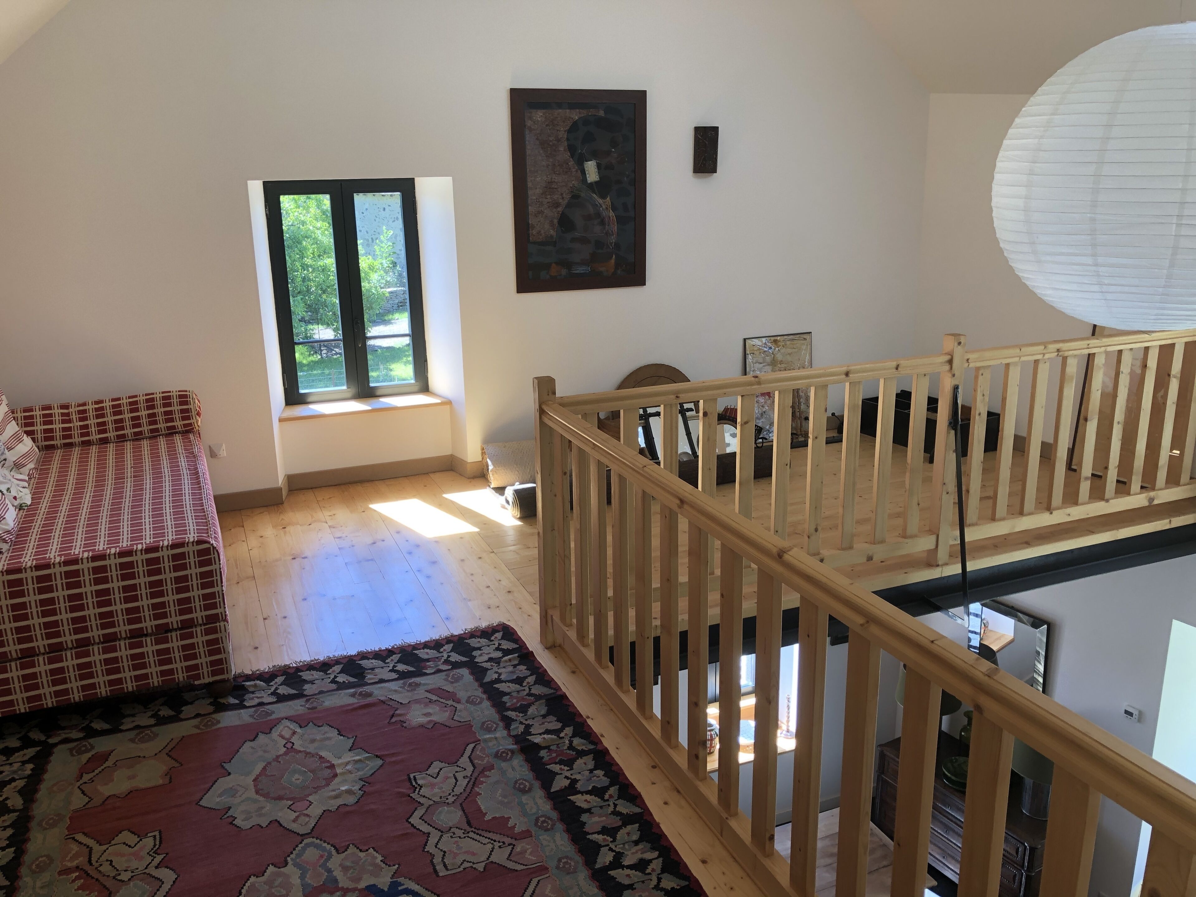 A room with a sofa, a patterned rug, a wooden railing, a window with a view, and a painting on the wall.