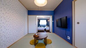 A room with tatami flooring, a low wooden table with cushions, a television on the wall, and a door leading to a bedroom with a bed and curtains. - Fuga-A (Fujikawaguchiko)