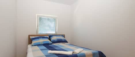 A single bed with a checkered bedspread, a window with blinds, and a plain white wall.