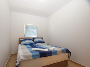 A single bed with a checkered bedspread, a window with blinds, and a plain white wall.