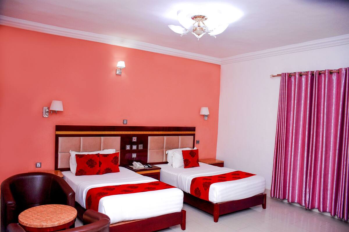 Une chambre d’hôtel avec deux lits, une tête de lit en bois, une chaise ronde en bois, une petite table ronde, un téléphone et un rideau.
