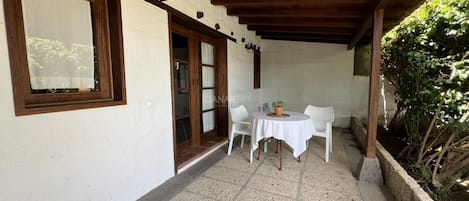 Un patio techado con mesa y sillas, techo de madera y pared blanca con ventana.