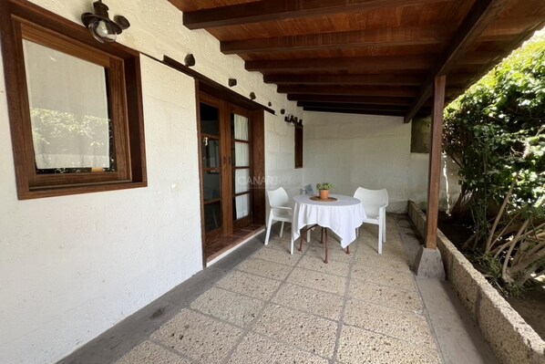 A covered patio with a table and chairs, a wooden ceiling, and a white wall with a window.
