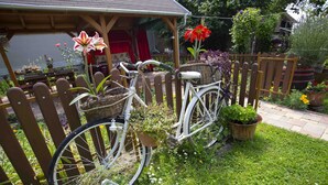 Una bicicleta blanca con cestas y plantas en macetas, estacionada en un jardín con una valla de madera y una casa al fondo.