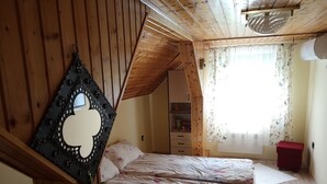 Un dormitorio acogedor con techo inclinado, paredes de madera, una cama con ropa de cama floral, un taburete pequeño rojo y una ventana con cortinas florales.
